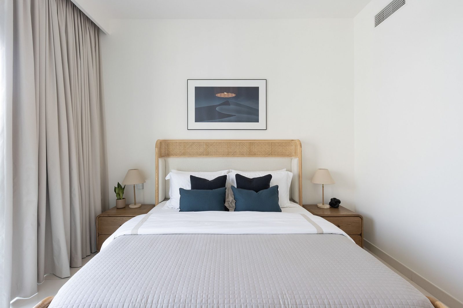Master bedroom furnished for long-term rent: rattan and ash bed, blue linen cushions, oak bedsides and a dune print