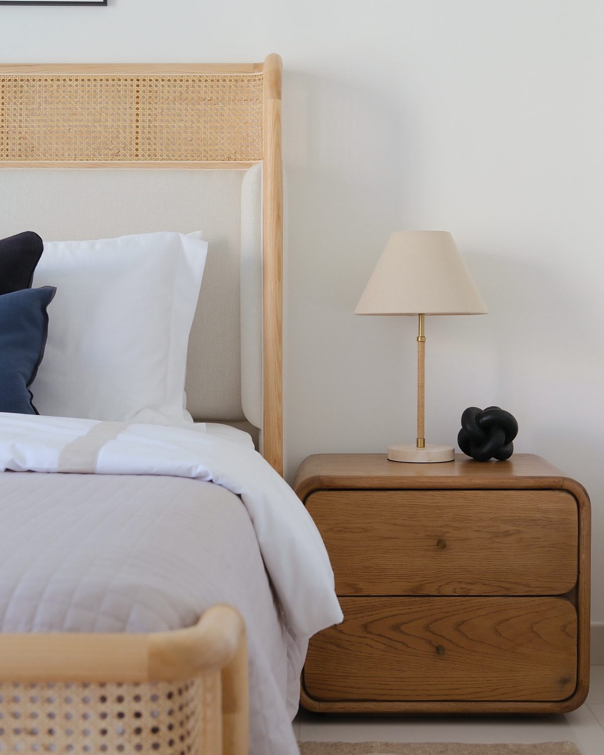 Bedside detail: rattan headboard, oak bedside table, jute-wrapped lamp and a black knot object