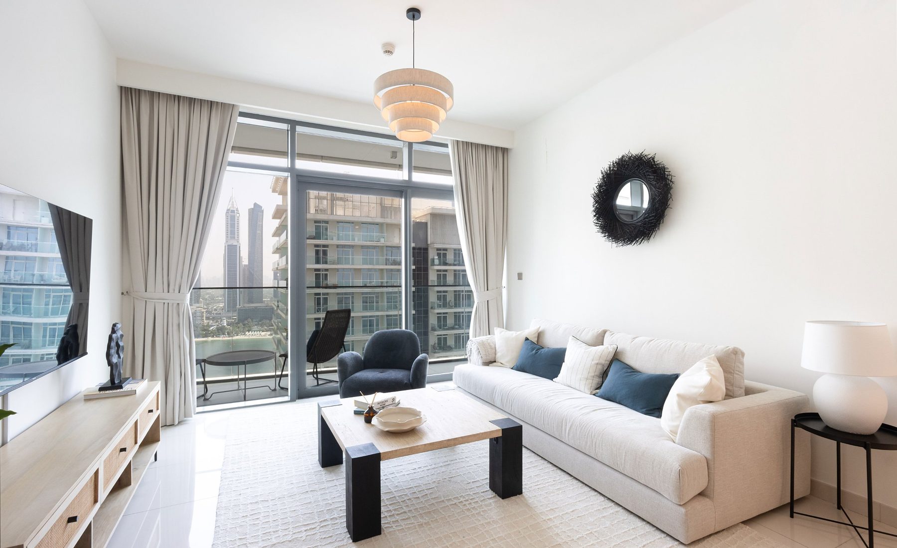 Furnished living room of a two-bedroom rental apartment in Dubai Harbour: linen sofa with blue cushions, navy armchair, travertine coffee table, black rattan mirror and the Marina through the window
