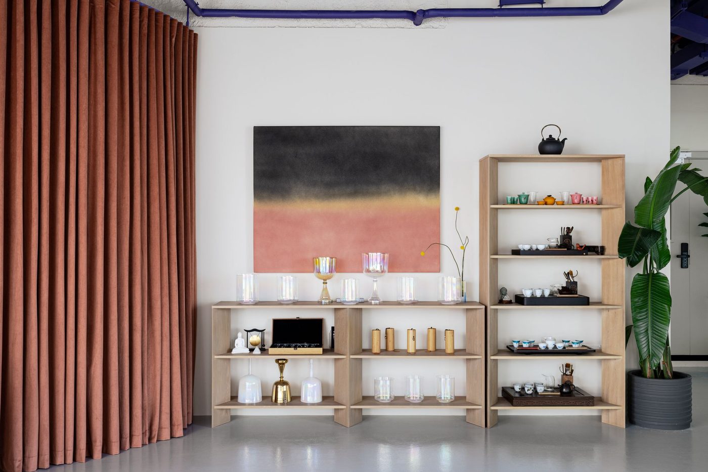 Wooden shelving with tea ceramics beside a rose curtain and abstract art