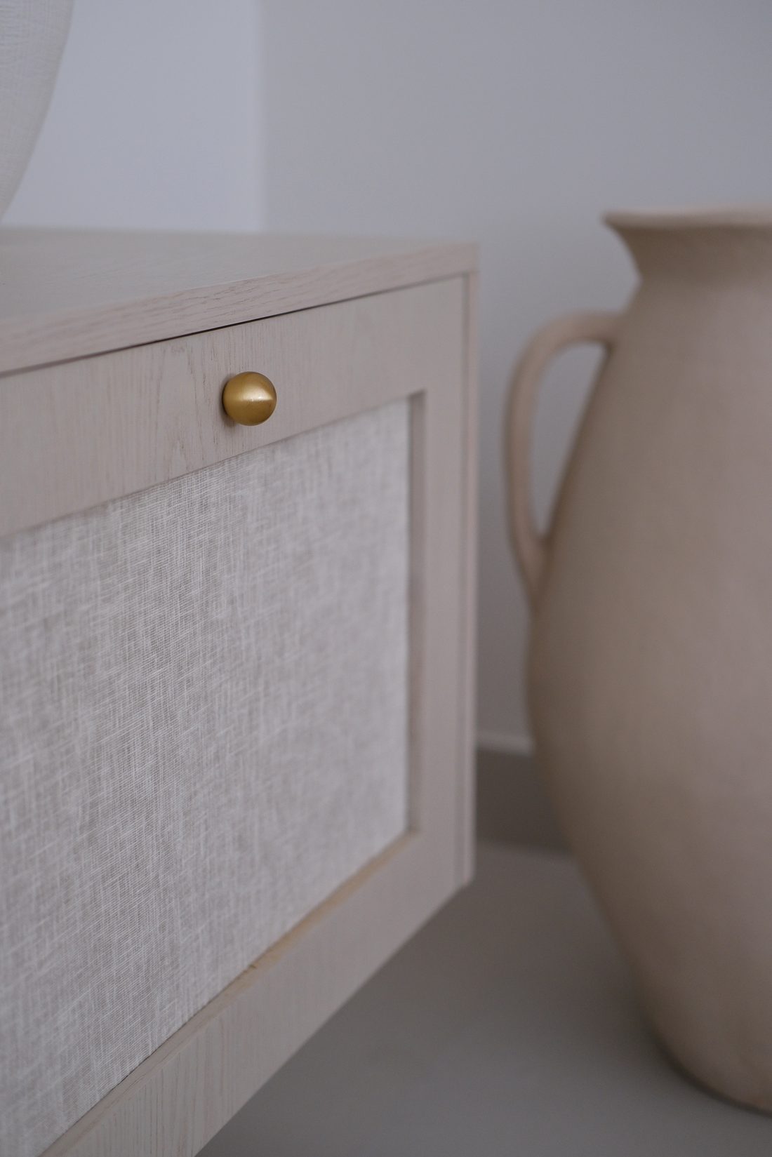 Bedside detail — limed oak and linen panel with a burnished brass knob beside a ceramic jug